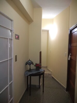 Um corredor estreito com uma porta à esquerda, uma pequena mesa com uma luminária e flores, e uma escada que leva a um andar superior.