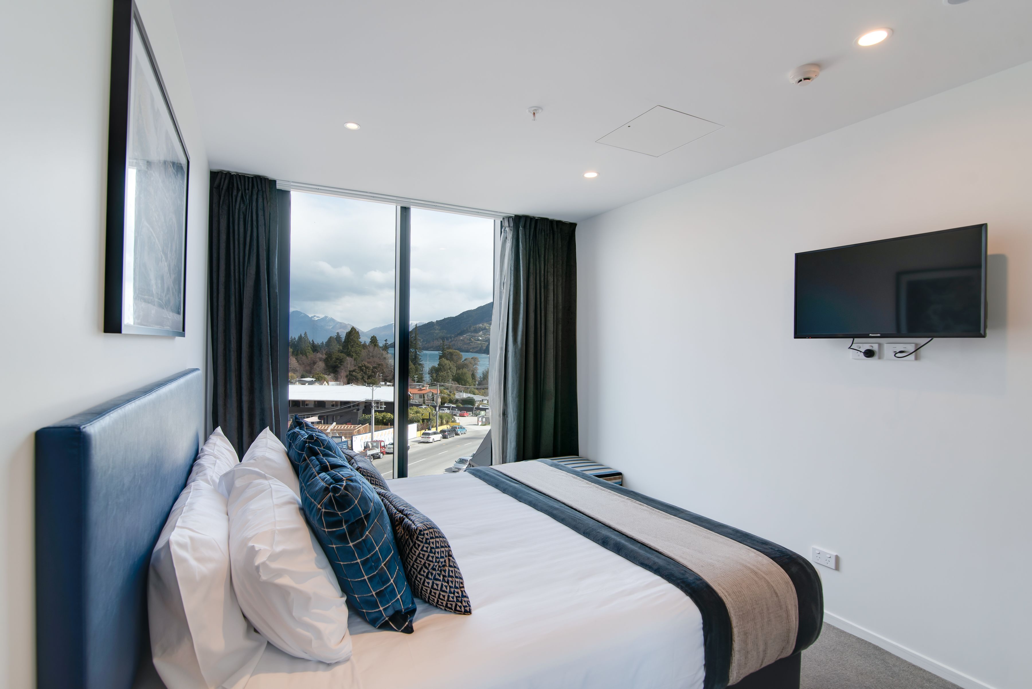 A hotel room with a large bed, a flat-screen TV mounted on the wall, and a view of a cityscape through the window.