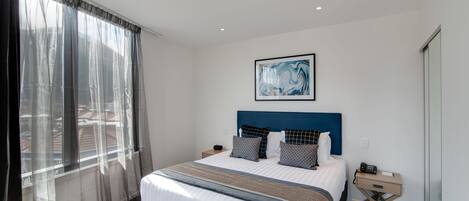 Une chambre d’hôtel moderne avec un grand lit, une table de chevet, une commode et une fenêtre avec des rideaux légers.