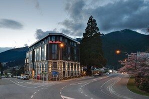 A modern building with a sign reading "RAMADA" at dusk, surrounded by mountains and a street with cars.