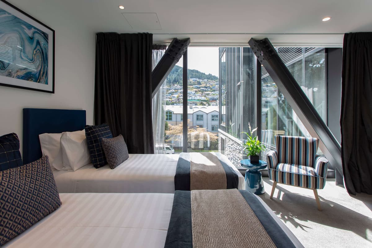 Ein Hotelzimmer mit zwei Betten, einem großen Fenster mit Vorhängen, Blick auf Gebäude und einem gestreiften Sessel.