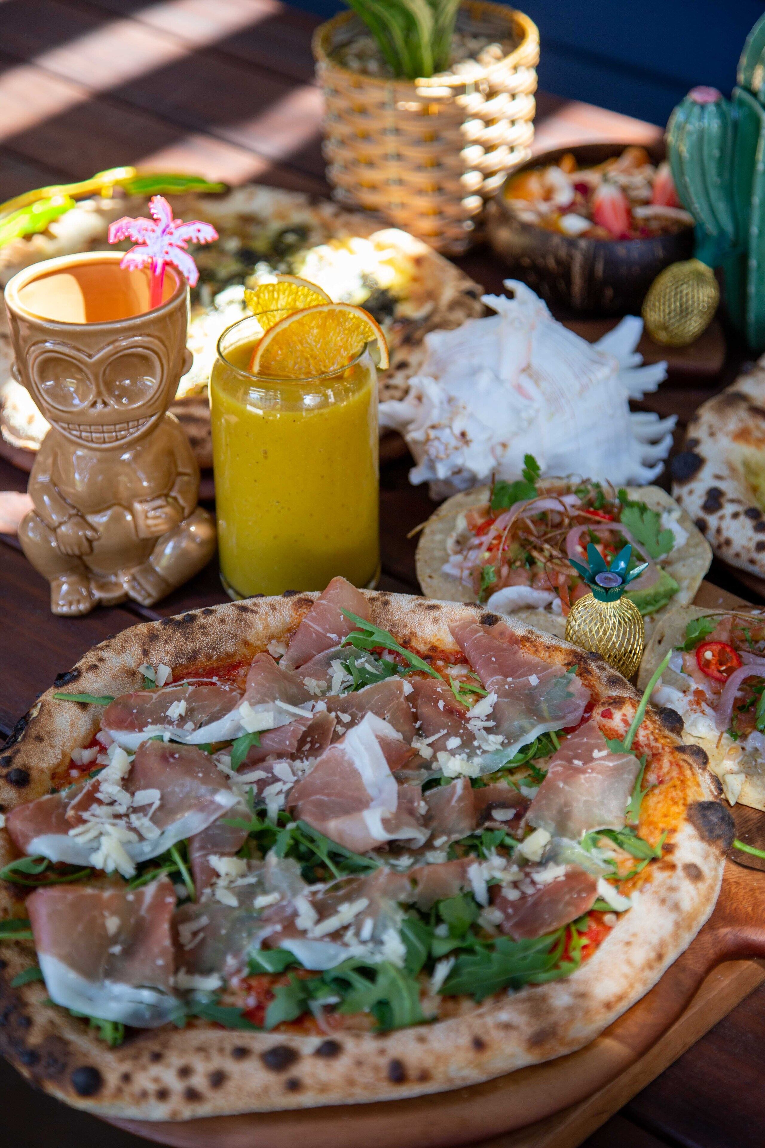 A variety of food items including pizzas and a drink, arranged on a wooden table.