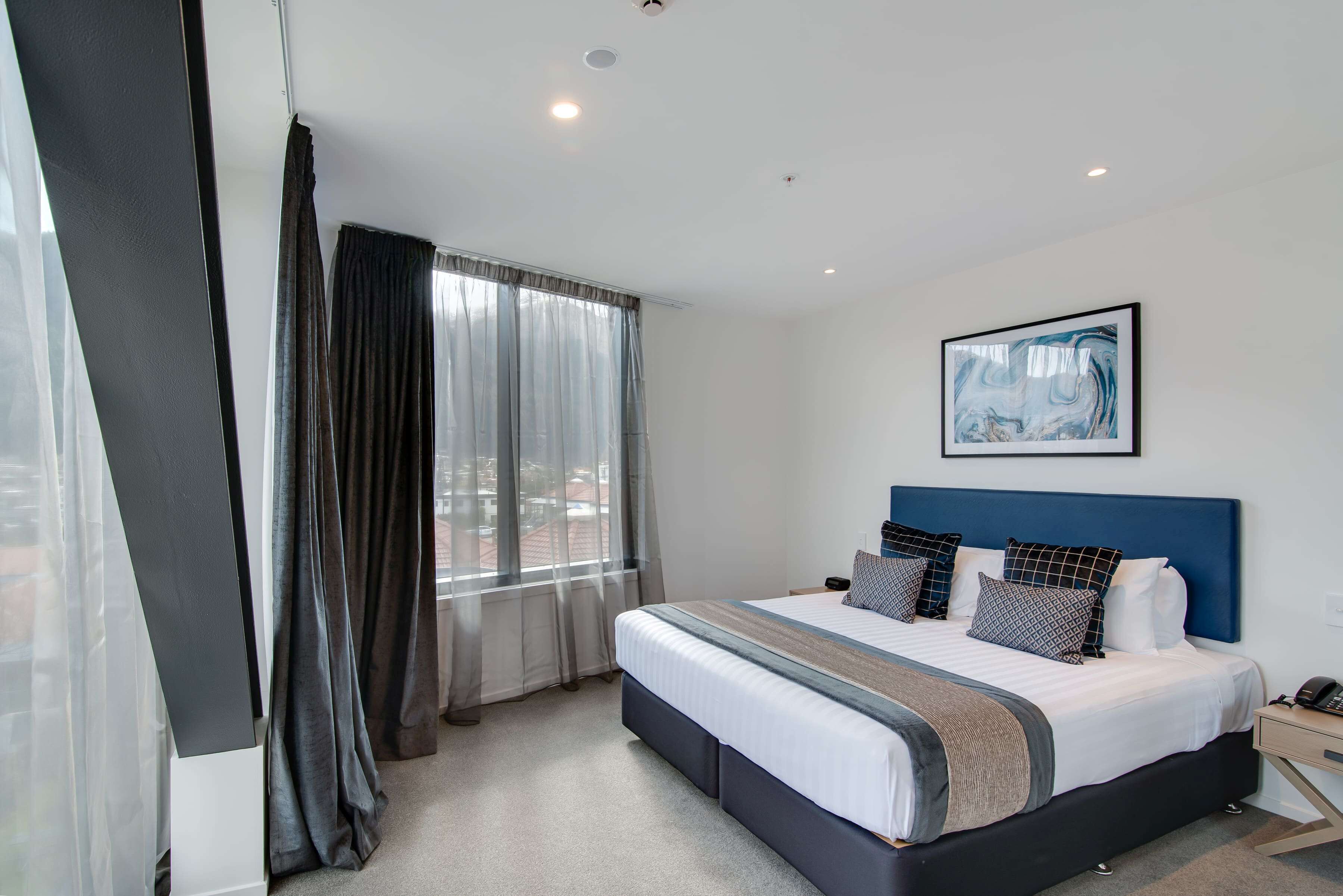 A modern hotel room with a large bed, a headboard, a bedside table, a framed artwork on the wall, and a window with curtains.