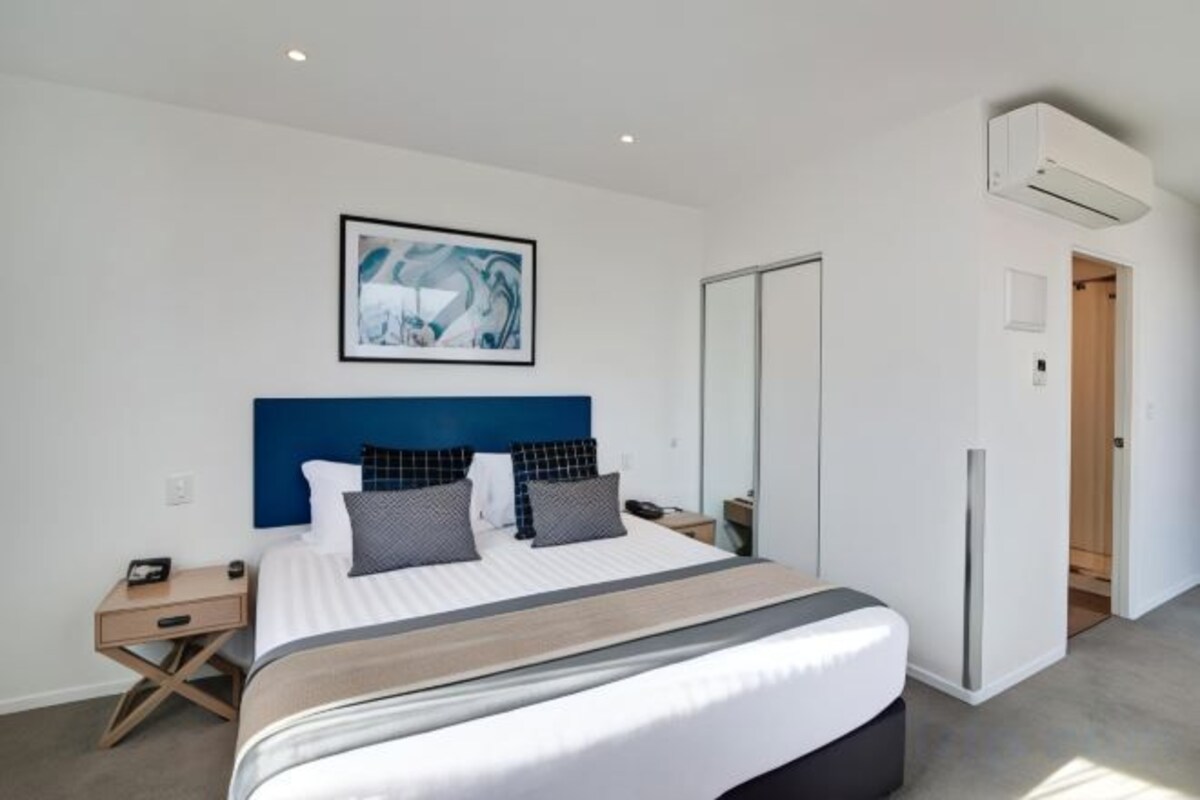 A modern bedroom with a large bed, a headboard, bedside tables, and a wall-mounted air conditioner.