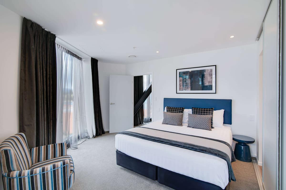 Ein modernes Hotelzimmer mit einem großen Bett, einem gestreiften Sessel, einem Nachttisch und Blick auf die Stadt durch das Fenster.