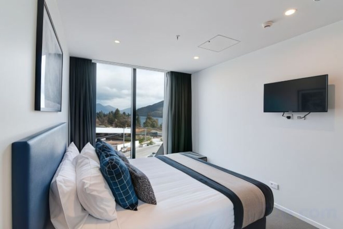 A hotel room with a bed, a TV mounted on the wall, and a large window offering a view of the outdoors.