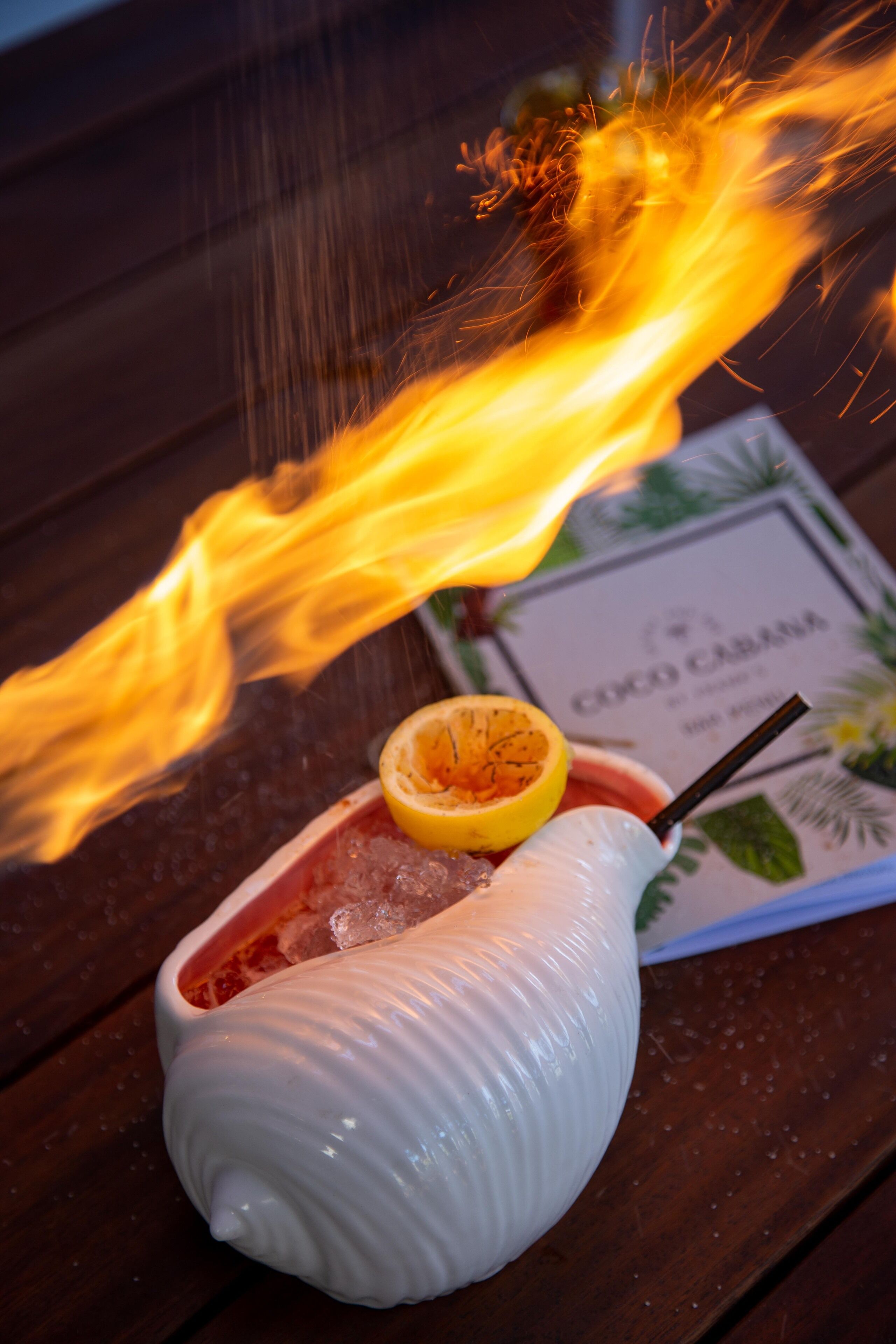 A flaming drink on a wooden table with a menu in the background.