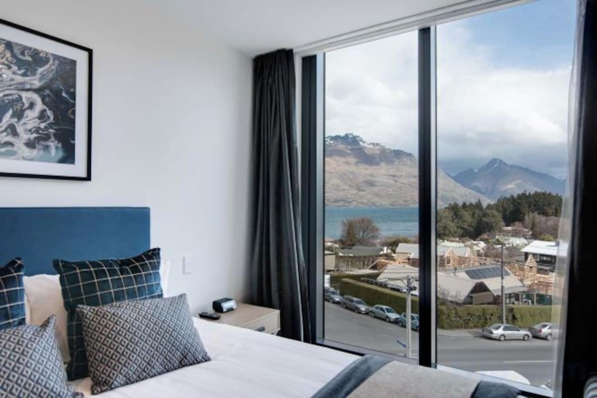 Un dormitorio con una cama grande, una mesita de noche y vistas a un pueblo y montañas a través de una ventana.