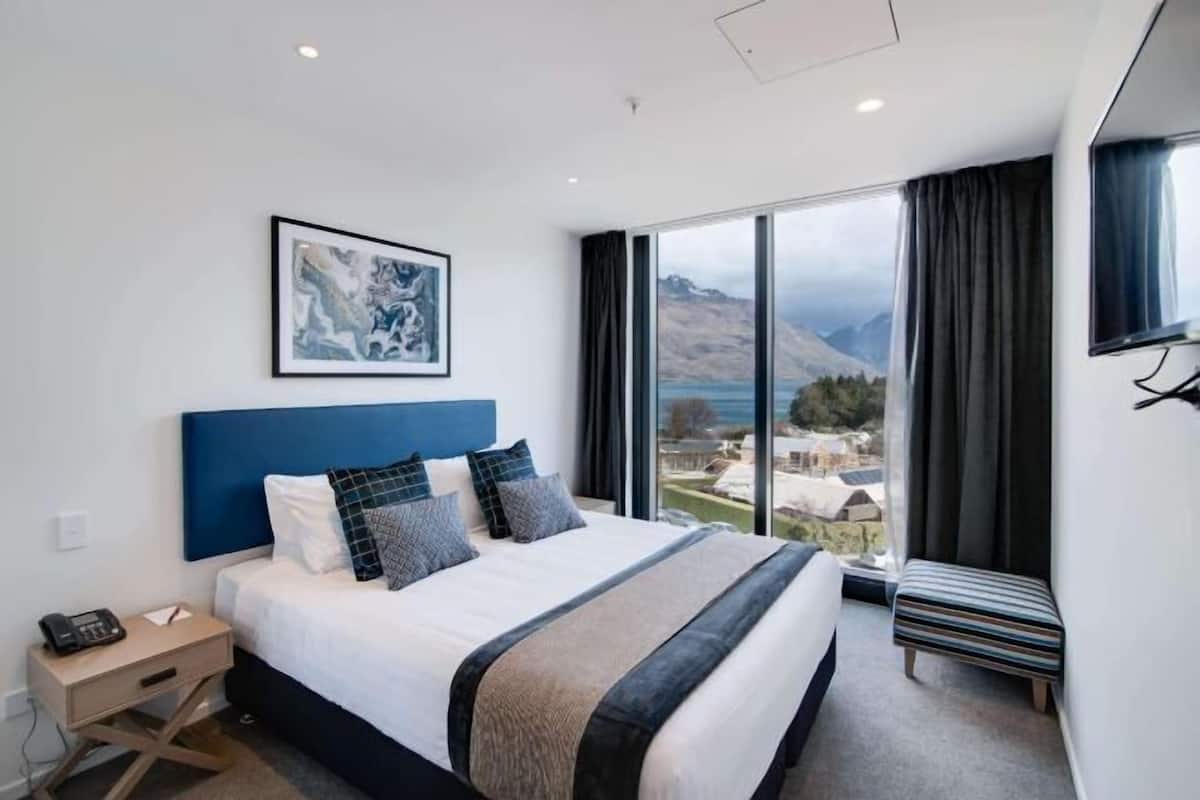 Ein modernes Hotelzimmer mit einem großen Bett, einem Nachttisch, einer Bank, einem Gemälde und Bergblick durch große Fenster.