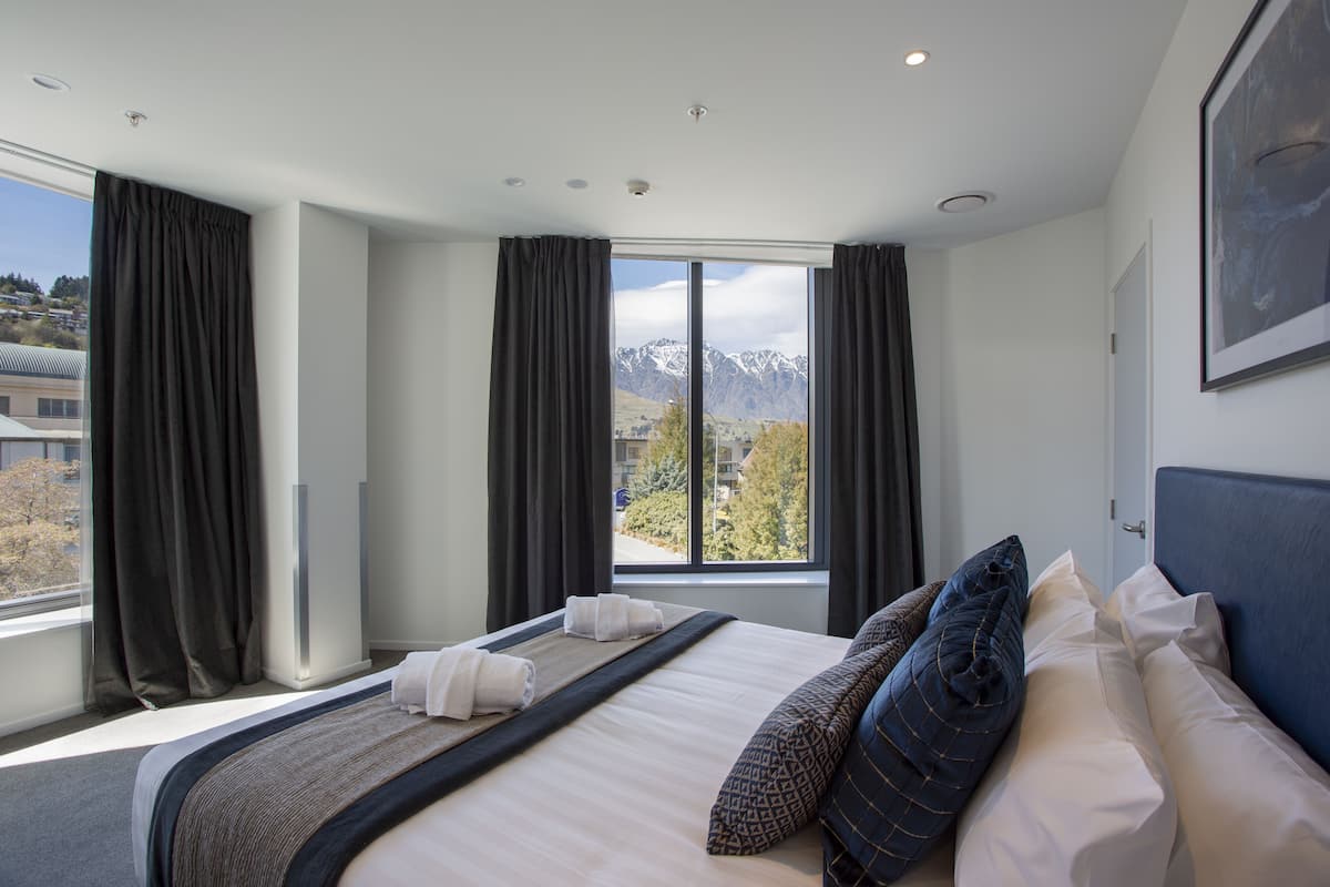 Ein Hotelzimmer mit einem großen Bett, dunklen Vorhängen, Blick auf Berge und einem gerahmten Bild an der Wand.