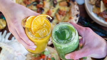 A person holding two drinks, one orange and one green, with a pizza and various food items in the background.