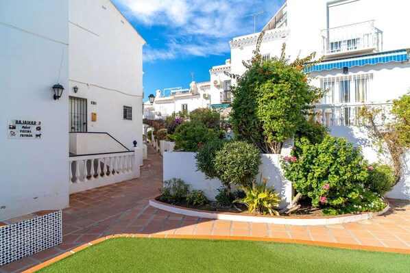 A narrow street with white buildings, a sign indicating a private pool, and well-maintained greenery.