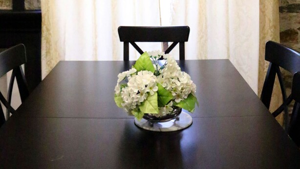 Una mesa de comedor con un jarrón de flores, sillas negras y una pared de piedra con ventana y cortinas.