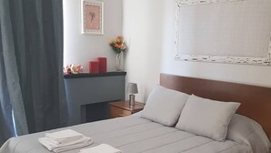 Una cama bien hecha con edredón gris y almohadas, una mesita de noche con lámpara, y un pequeño florero con flores en la pared.