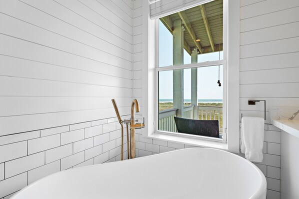 A modern bathroom with a freestanding bathtub, a window with a view of the beach, and white tiled walls.