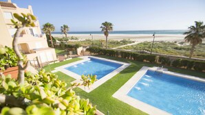 Zona de piscina con dos piscinas rectangulares, rodeada de césped cuidado y palmeras, con vistas a una playa.