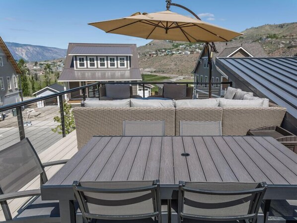 Une terrasse en bois avec une table et des chaises, un grand parasol et un canapé en osier agrémenté de coussins.