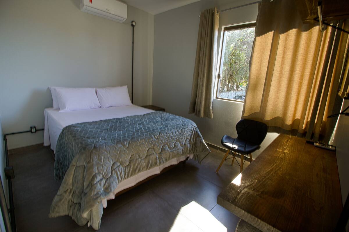 Un dormitorio con cama, una silla, un ventanal con cortinas y un escritorio de madera.