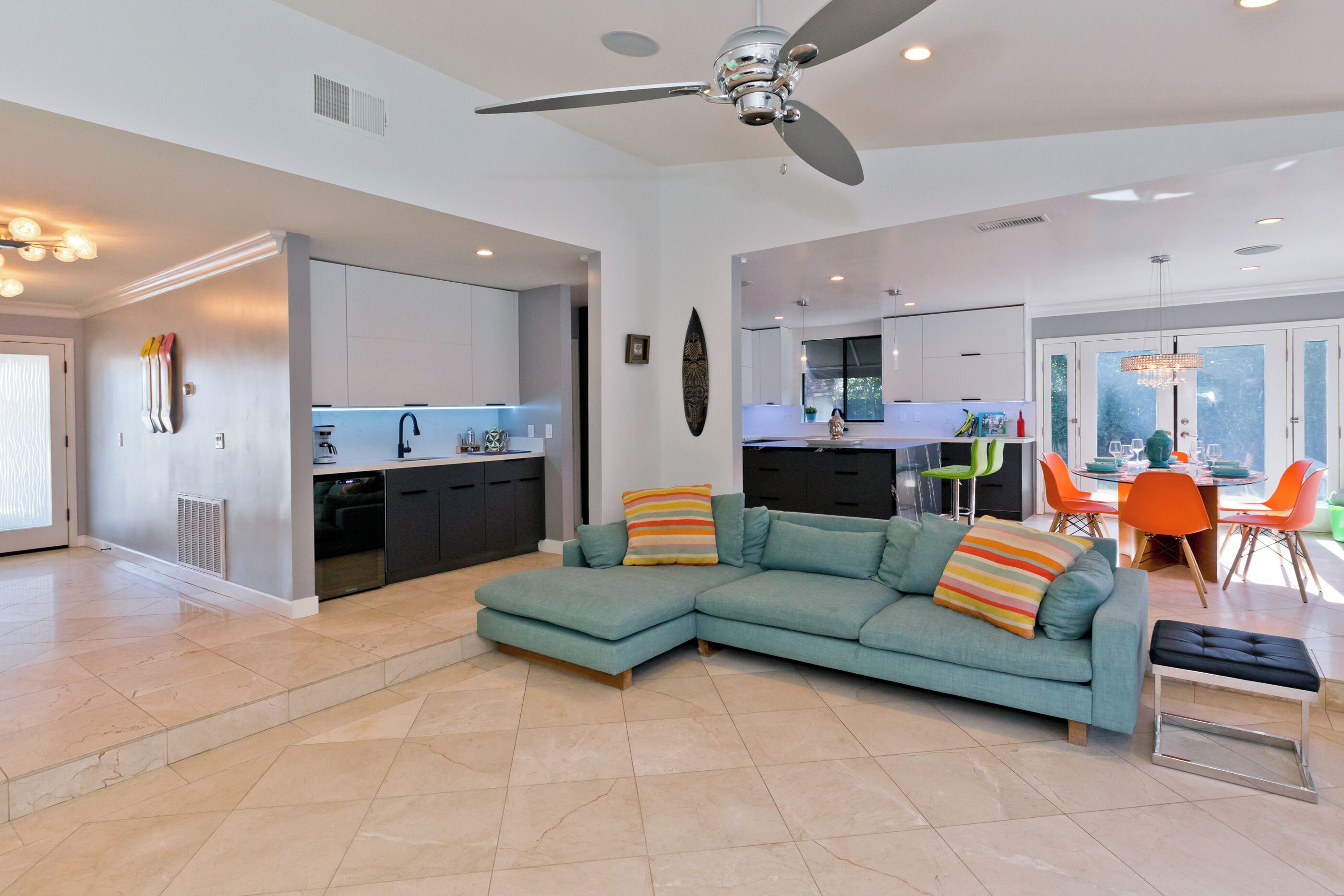 A modern living room with a teal sectional sofa, a dining area with orange chairs, and a kitchen in the background.