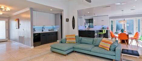 A modern living room with a teal sectional sofa, a dining area with orange chairs, and a kitchen in the background.
