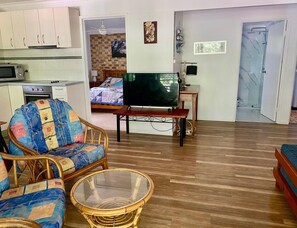 Un salon comprenant un téléviseur, un fauteuil en osier, une table basse en verre, un lit avec une courtepointe et un coin cuisine.
