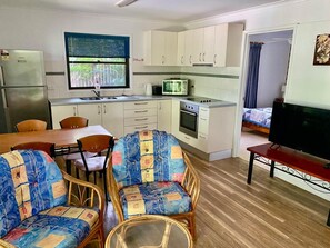 A compact kitchen with a microwave, oven, and sink, a dining area with a table and chairs, and a television in a bedroom. - Spacious Straddie family retreat sleeps up to 10, walk to Cylinder Beach (North Stradbroke Island)