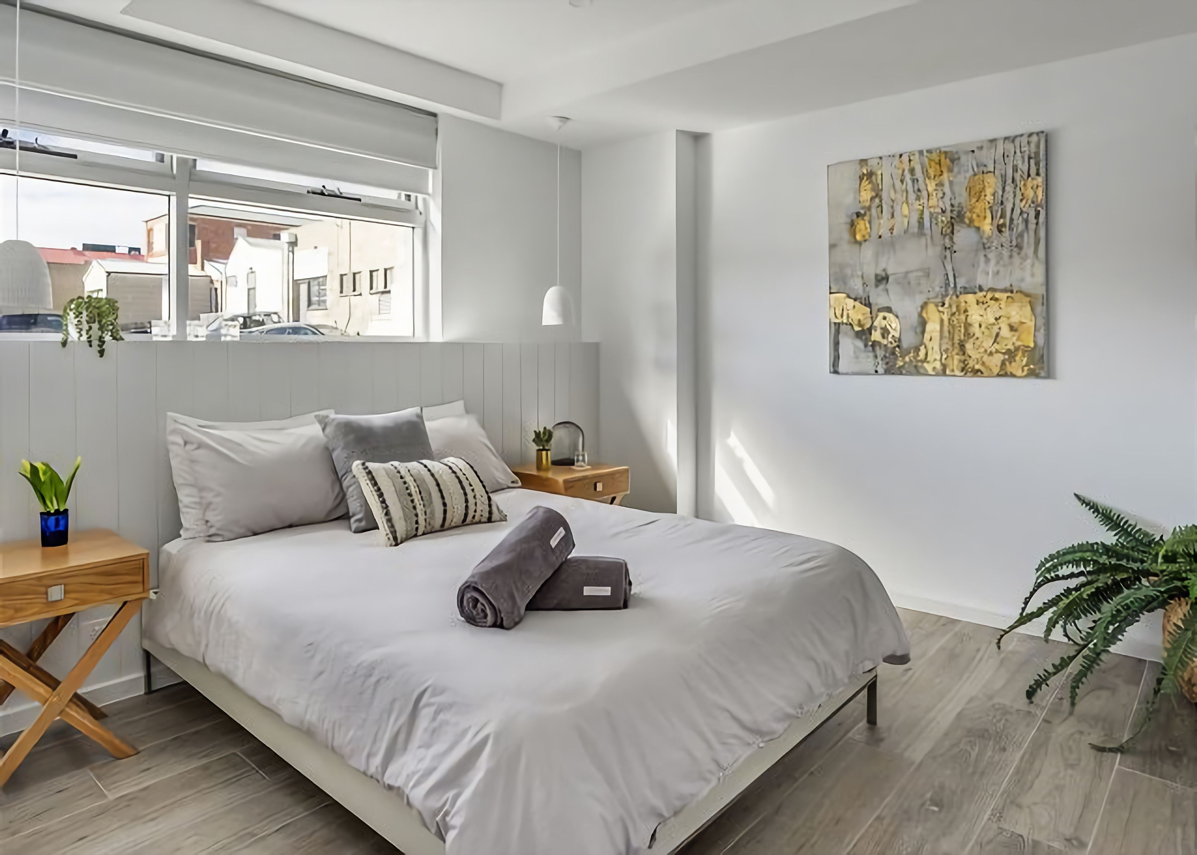 A modern bedroom with a large bed, a bedside table, a nightstand, a window, and a painting on the wall.