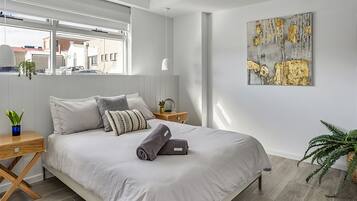 A modern bedroom with a large bed, a bedside table, a nightstand, a window, and a painting on the wall.