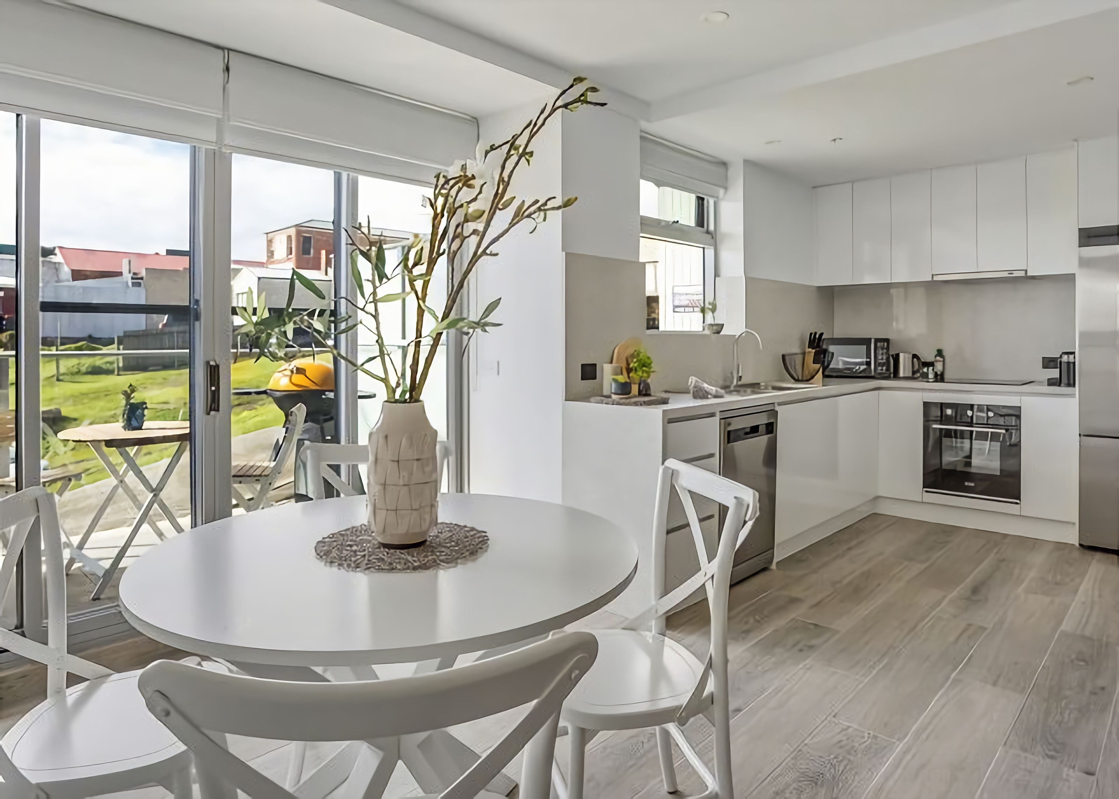 A modern kitchen with white cabinetry, a dining area with a round table and chairs, and a view of the outdoors through large windows.