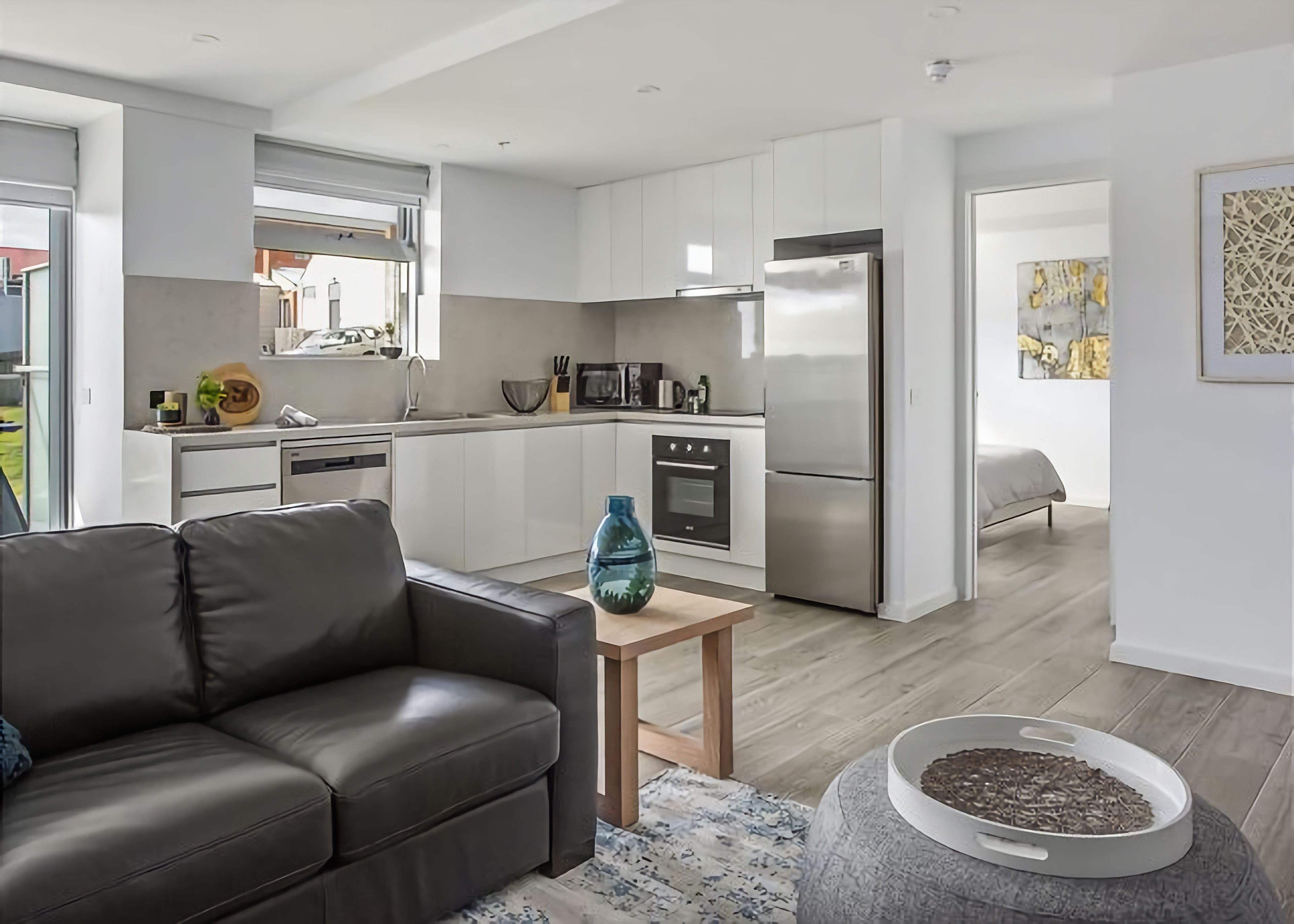 A modern kitchen with white cabinets, a stainless steel refrigerator, and a dining area with a leather sofa and a small table.