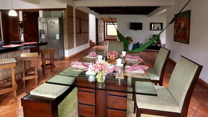 A dining area with a long table set for six, surrounded by wooden chairs with cushioned seats. A green hammock is visible in the background, and a television is mounted on the wall. - Casa de las flores (Villa de Leyva)