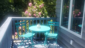 Una terraza de madera con una mesa y sillas turquesas, rodeada de exuberante follaje y flores rosadas.