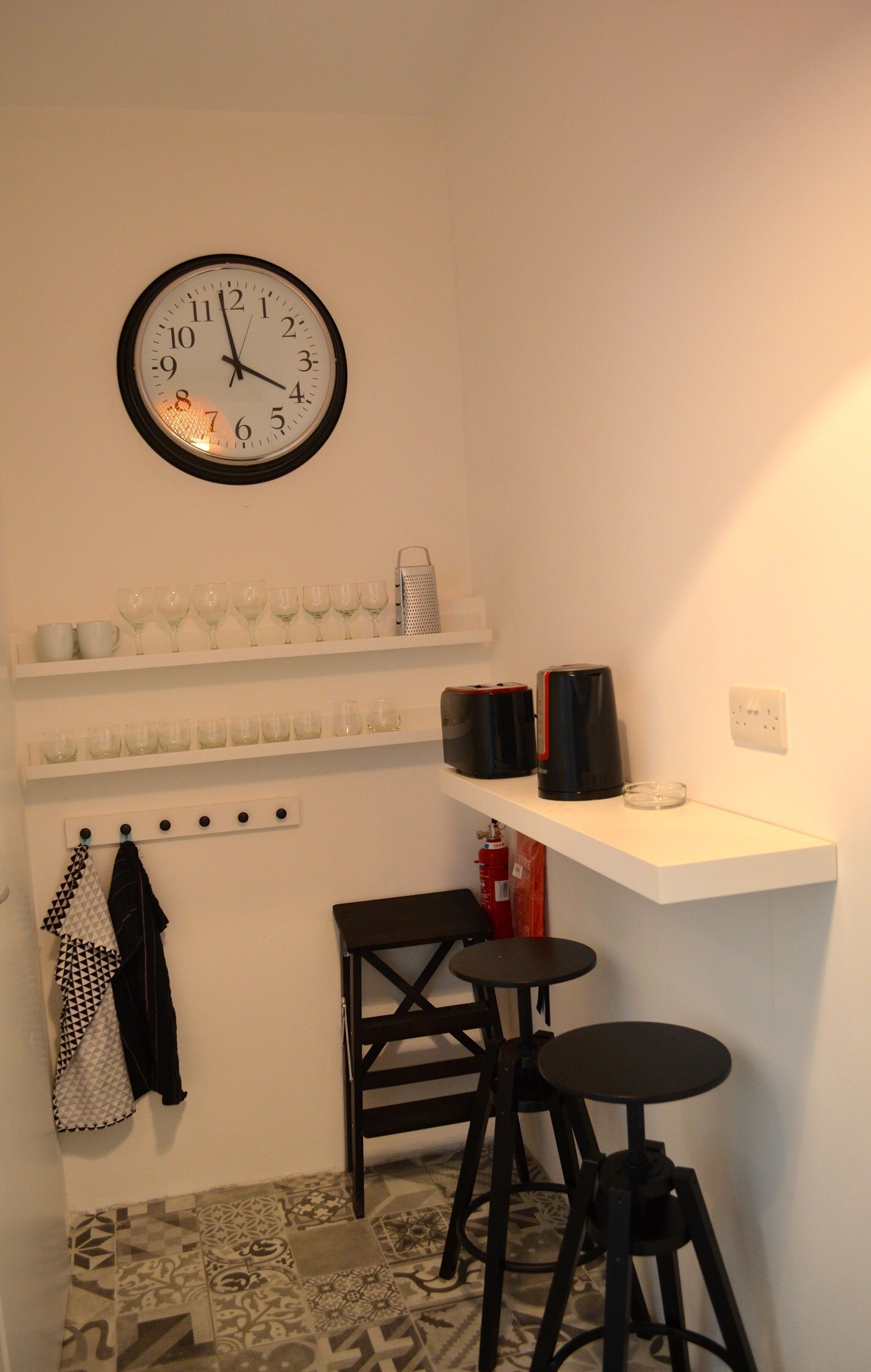 A small kitchen with a clock, shelves with glasses, a coffee maker, and two stools.