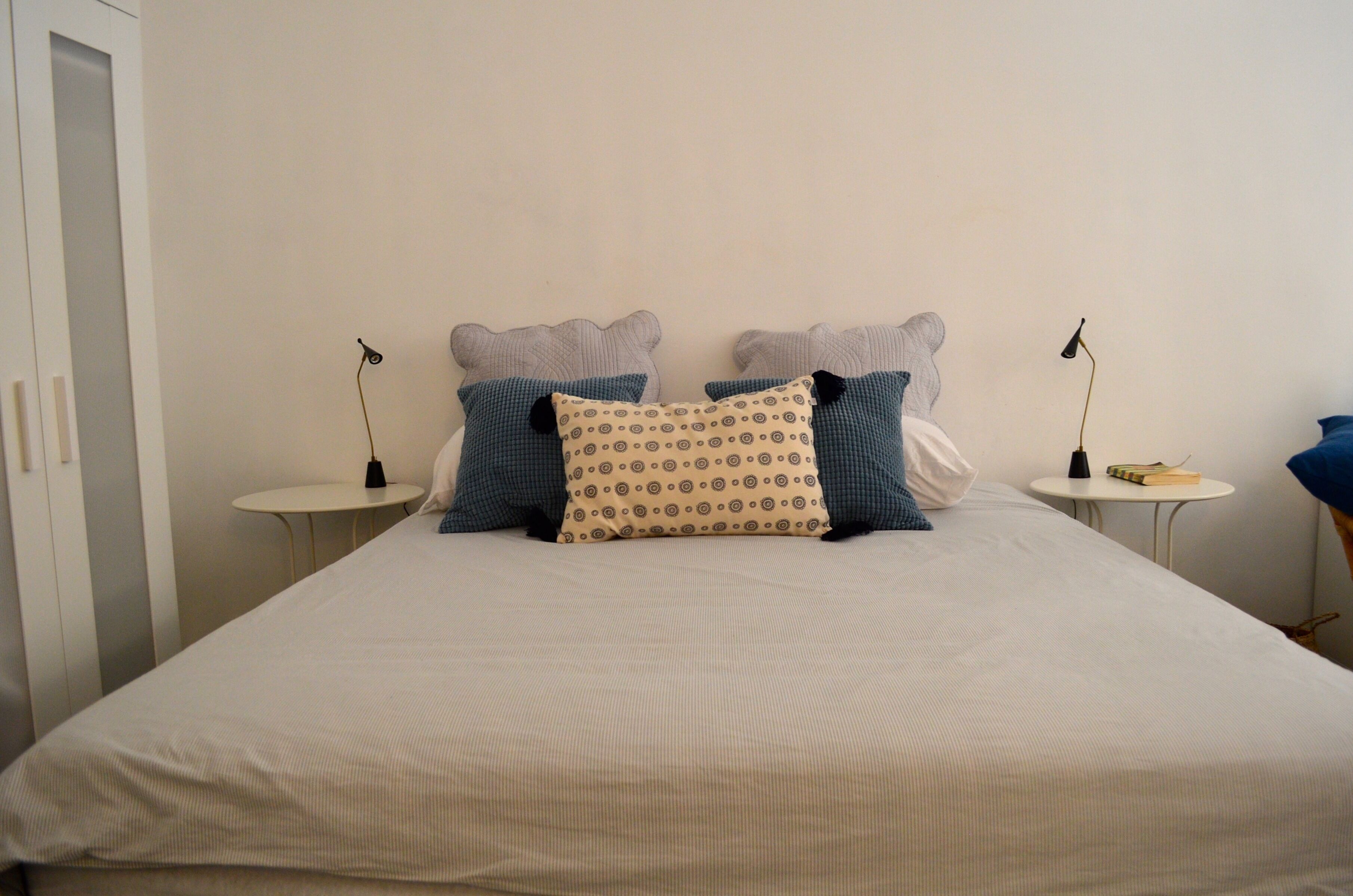 A neatly made bed with a patterned pillow, two bedside tables with lamps, and a small table with a book and a lamp.