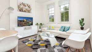 A modern living room with a flat-screen TV, white furniture, a glass coffee table, and a view of greenery outside. - Stunning Sydney Home 6 (Millers Point)