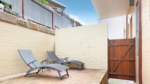 Un patio con dos sillones de descanso, piso de ladrillo, un portón de madera y una pared blanca.