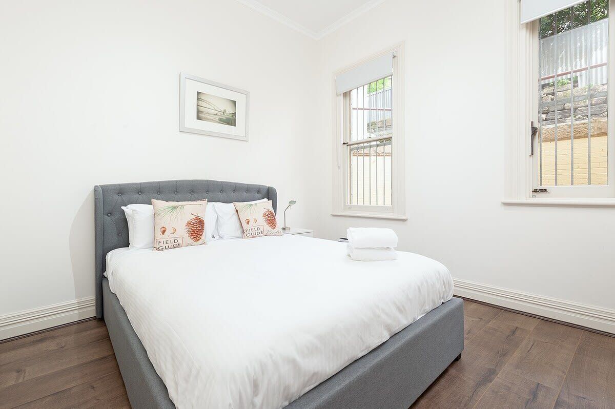 A neatly made bed with a grey headboard, white bedding, and decorative pillows. There is a framed picture on the wall and a window with bars.