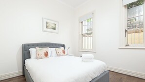 Una cama bien hecha con un cabecero gris, ropa de cama blanca y almohadas decorativas. Hay un cuadro en la pared y una ventana con barrotes.