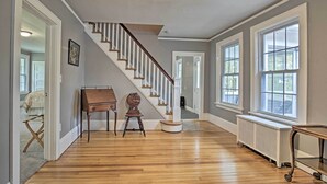 Una habitación amplia con pisos de madera, una escalera con barandales blancos, ventanas grandes y un radiador.
