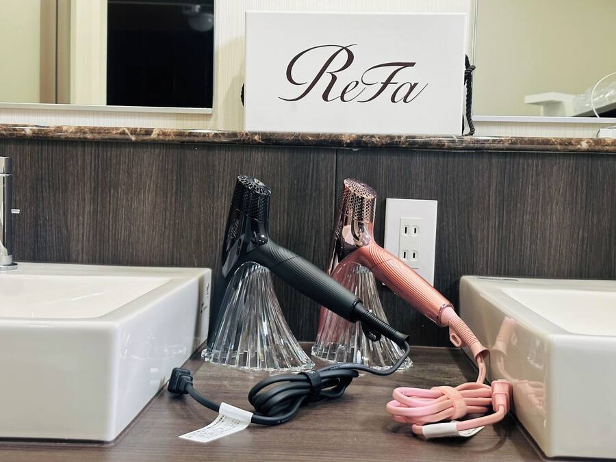 A bathroom with a mirror, a ReFa sign, two hair dryers, and a power outlet.