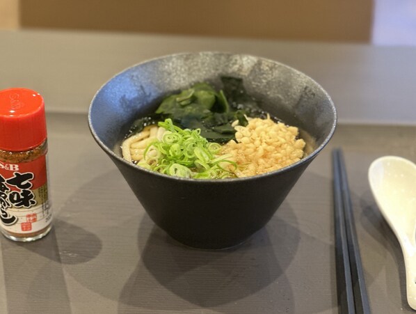 Un tazón de ramen con cebolletas y alga, una botella roja de condimento y palillos sobre una mesa.