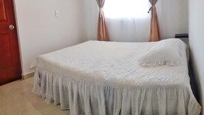 A single bed with a white bedspread and a ruffled skirt, a wooden door, and a window with curtains. - Come to Rionegro, Come to Life (Rionegro)