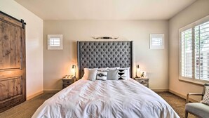 Een slaapkamer met een gestoffeerd hoofdeinde, een groot bed met wit beddengoed, twee nachtkastjes met lampen en een raam met jaloezieën.
