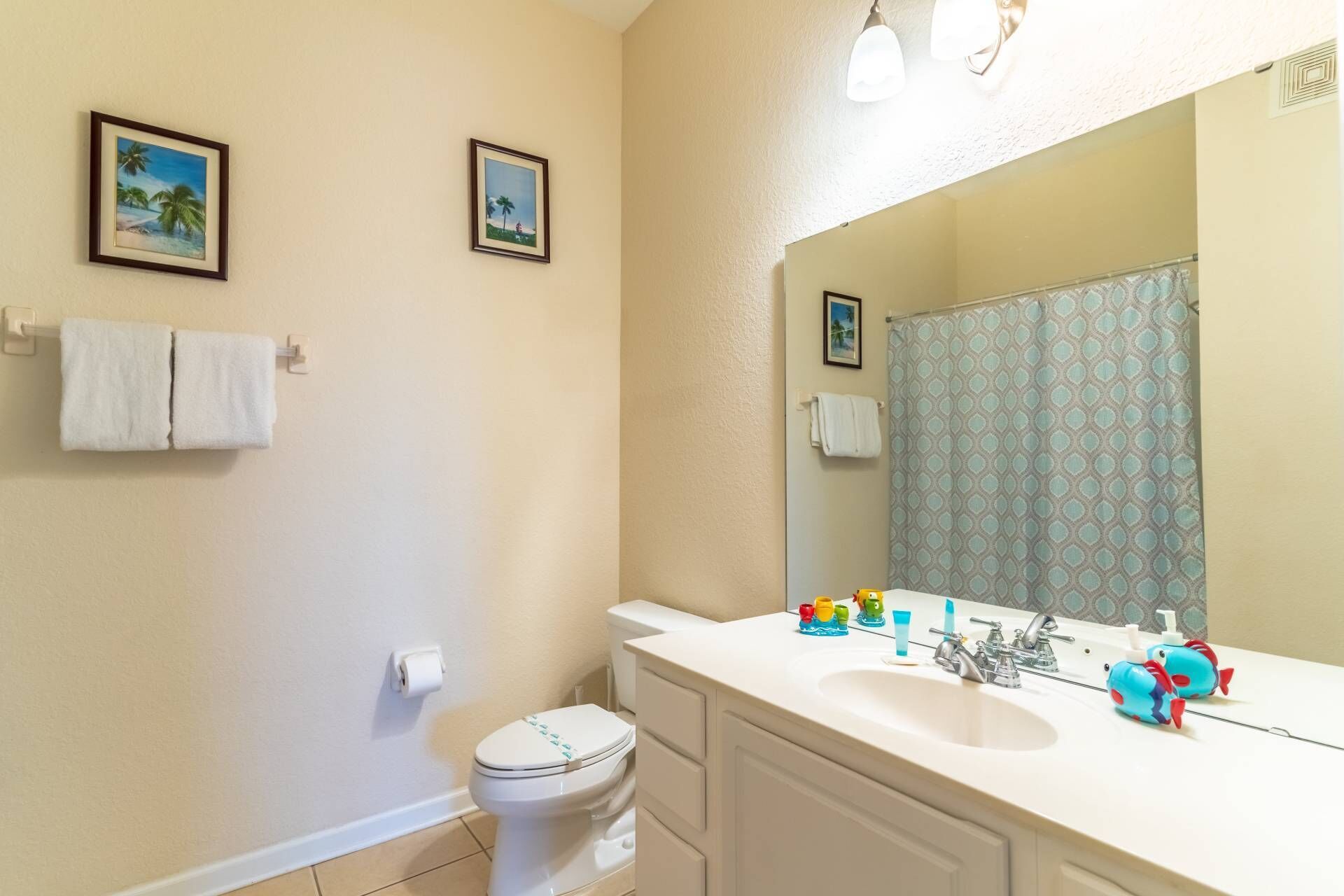 Une salle de bain avec des toilettes, un lavabo et un miroir. Deux tableaux encadrés sont accrochés au mur, ainsi qu’un porte-serviettes avec deux serviettes et un rideau de douche à motifs géométriques.