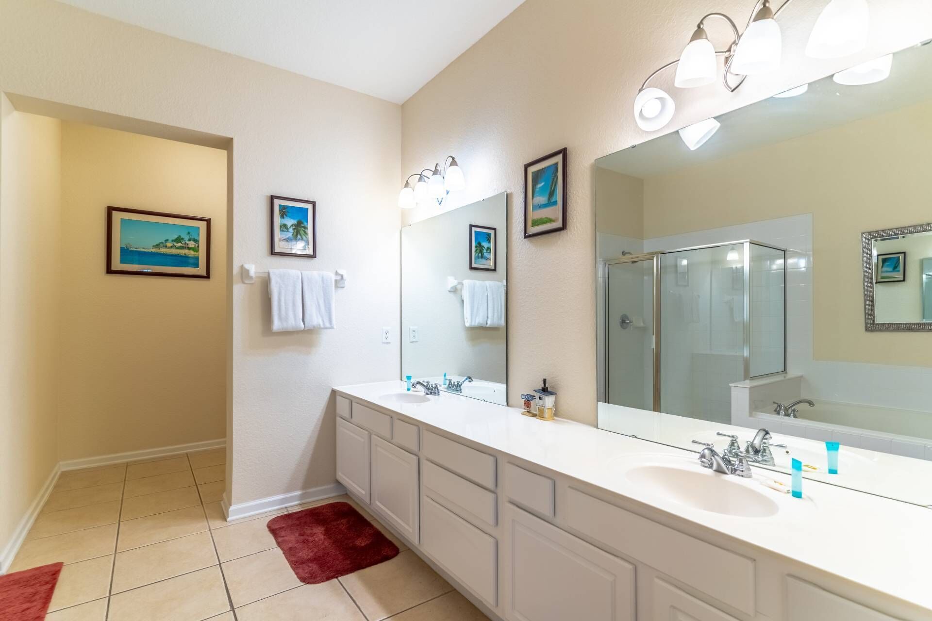 A bathroom with a large mirror, two sinks, and a shower. There are towels hanging on the wall and a red rug on the floor.