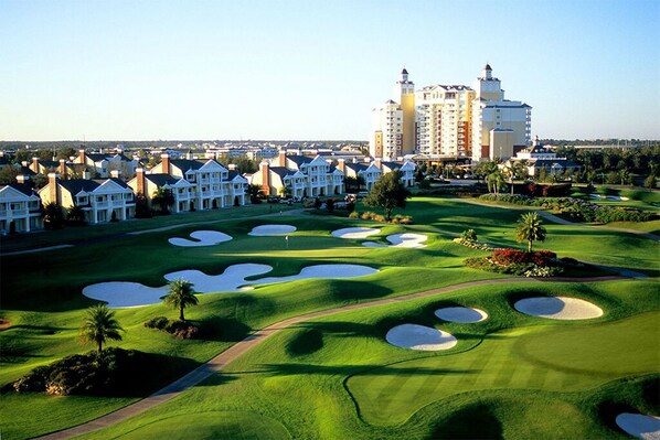A golf course with bunkers, a clubhouse, and a hotel in the background.