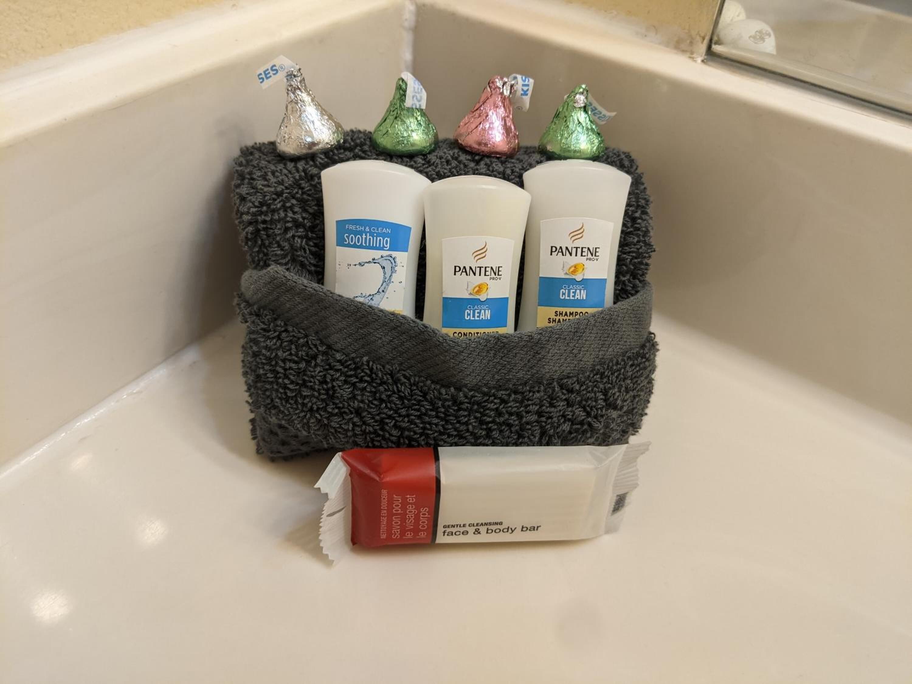 A bathroom sink with a black towel holder containing three bottles of Pantene Pro-V Classic Clean shampoo and conditioner, a bar of gentle cleansing face and body soap, and four wrapped chocolates.