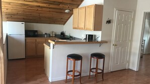 A kitchen with wooden cabinets, a white refrigerator, a microwave, and a coffee maker. There are two wooden stools and a white door. - Misty Lake Carriage House (Wasilla)