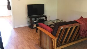 A cozy living room with a wooden ceiling, a flat-screen TV on a stand, a wooden sofa with cushions, and a glass coffee table. - Misty Lake Carriage House (Wasilla)