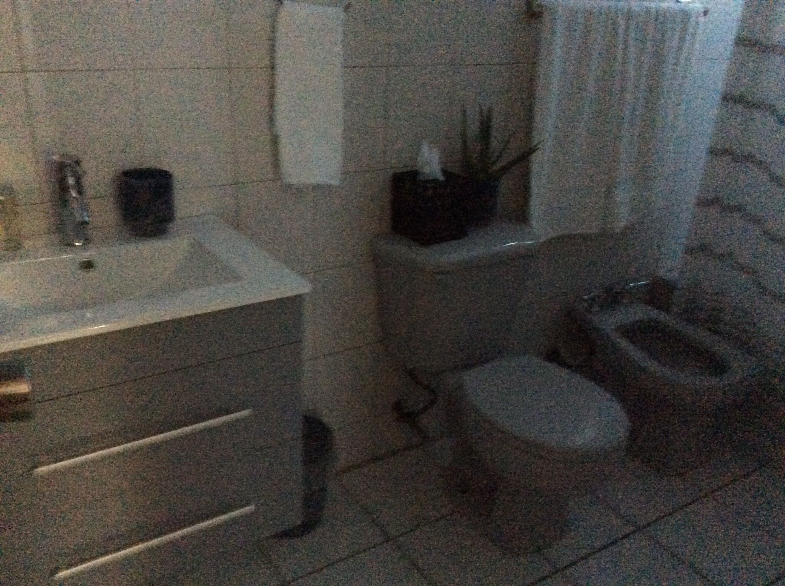 Un baño con lavamanos, inodoro y bidé blancos. Hay una toalla colgada en la pared y un pequeño cubo de basura.